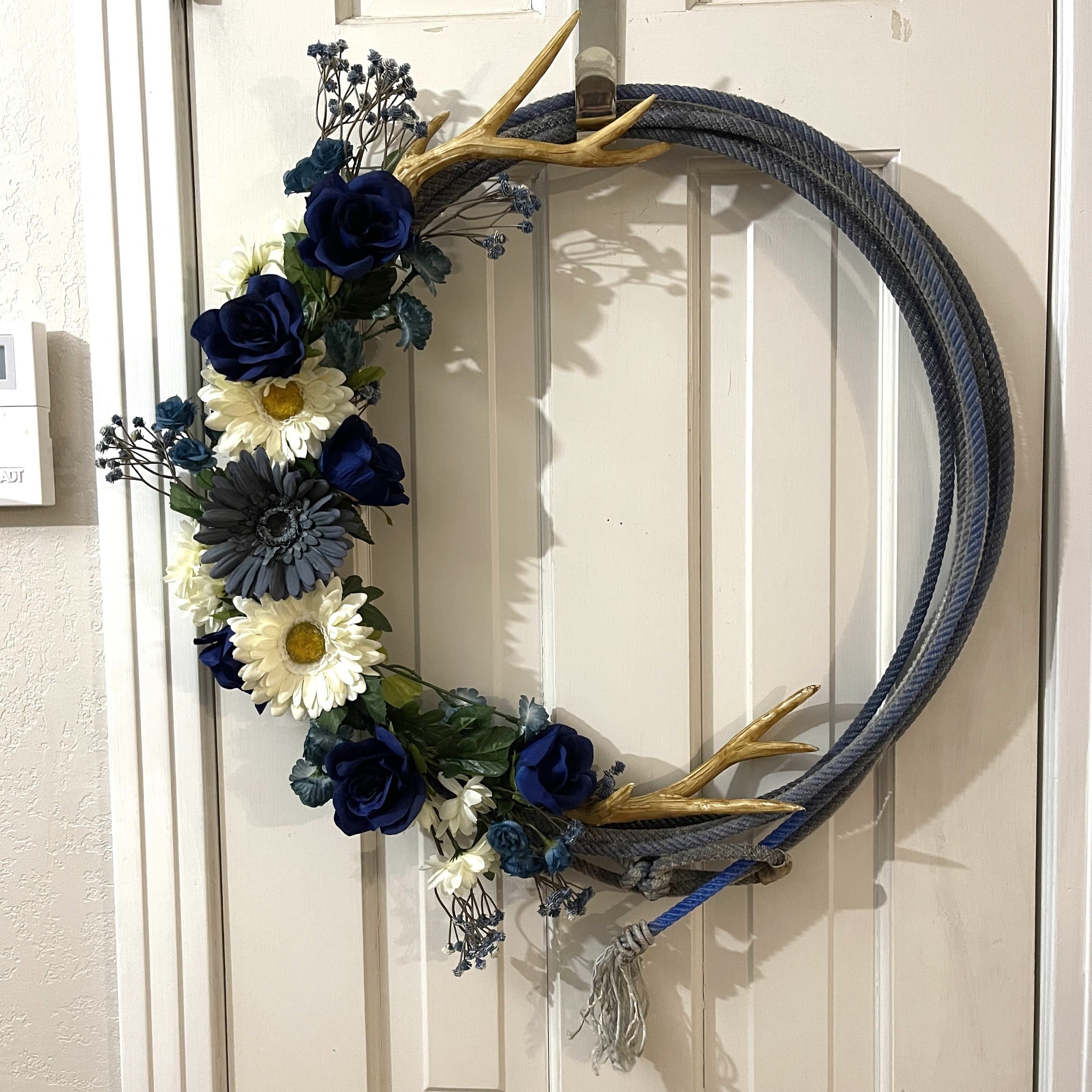 Handcrafted wreath on an upcycled lariat roping rope. Width: 21 inches. Florals: Navy blue and white gerbera daisies, navy blue gypsophila, royal blue roses, navy blue tea roses and pom poms, white daisies, and plastic antlers. 